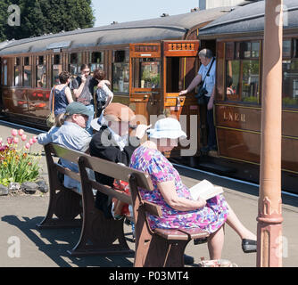 Busy platform scene at vintage railway station in summer sunshine. Passengers on & off vintage train. British folk sat on bench waiting & enjoying sun. Stock Photo