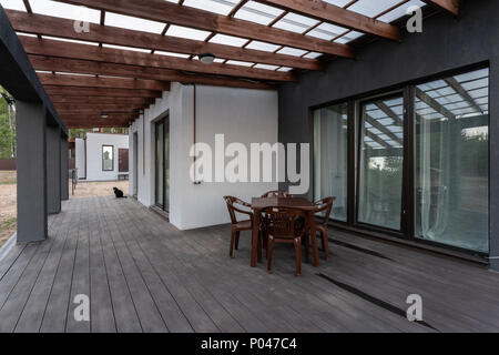Side view of an open veranda in front of a modern forest cottage. Pine young forest under the sunset rays in the background. Stock Photo