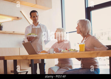 Top service. Pleasant young waiter serving a salad to two senior businesswomen sitting in cafe and discussing their new project Stock Photo