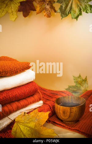 Stack of bright warm knitwears, colorful maple leaves and wooden cup of hot tea on light background. Stock Photo