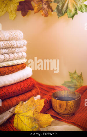 High stack of warm knitwears, maple leaves and wooden cup of hot tea on light beige background. Stock Photo