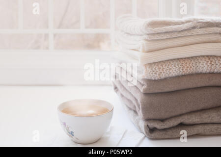 Stack of warm knitwears and cup of hot tea on windowsill on white window background. Stock Photo