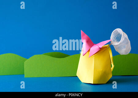 Paper backpack with butterfly net on grass field at blue background. Summer adventure. Camping and hiking or tourism concept. Origami. Paper cut. Stock Photo