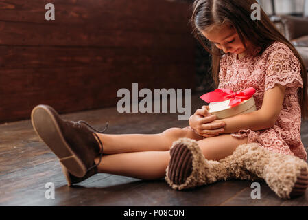 adorable little child holding giftbox in shape of heart while sitting on floor Stock Photo