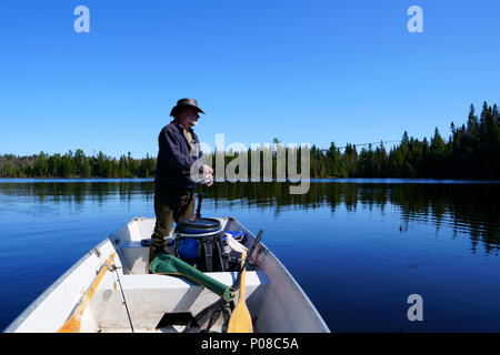 A peaceful caucasian man, 50's, wearing a white beard, a leather hat Australian style and a blue coat, is fishing in his boat on a lake with blue sky. Stock Photo