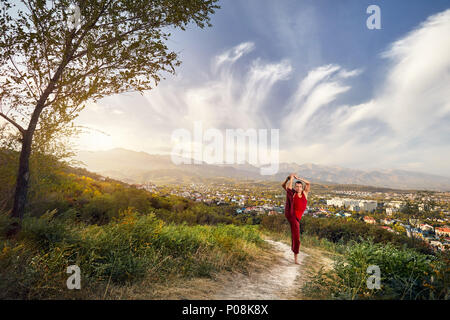 Fit man in red costume doing yoga leg split asana in the park with city and mountain on background in Kazakhstan Stock Photo