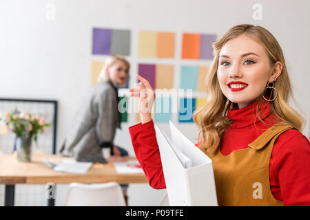 smiling magazine editor with folder in modern office with colleague and color palette on wall behind Stock Photo