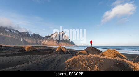 Man in red jacket standing on dunes with dry grass, black lava beach, sandy beach, mountains Klifatindur Stock Photo