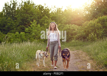 Happy young woman walking her dogs along a grassy rural track in spring looking at the camera Stock Photo