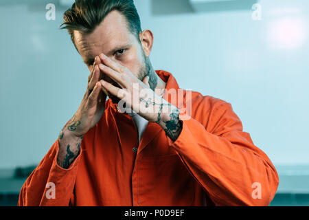 prisoner with tattoos covering face with hands and looking at camera Stock Photo