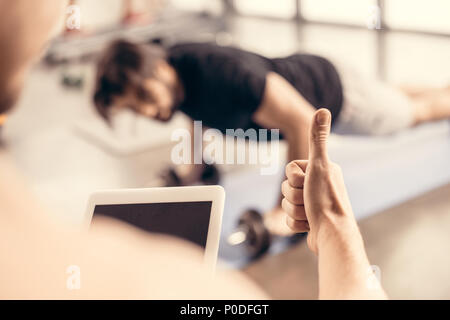 trainer holding tablet and showing thumb up to sportsman doing push ups on dumbbells in gym Stock Photo