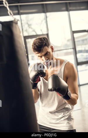 concentrated young sportsman in boxing gloves looking at punching bag in gym Stock Photo