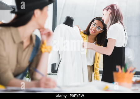 Cropped shot of young woman drawing sketch and looking at smiling colleagues working with dress on dummy Stock Photo