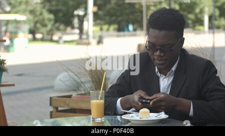 Relaxed carefree young afro american man in stylish eyewear sitting alone at cafe table, using smart phone, reading text message with joyful smile during lunch Stock Photo