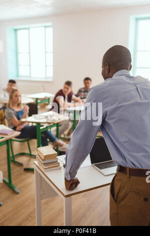 Rear view of teacher at table with laptop talking to pupils in classroom Stock Photo