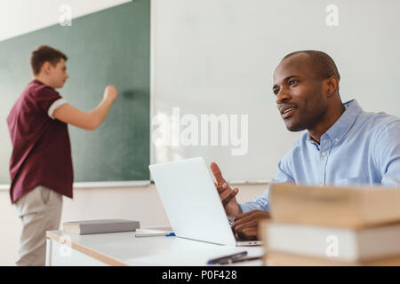 High school teenage student writing on chalk board and african american teacher sitting at desk with laptop and explaining Stock Photo