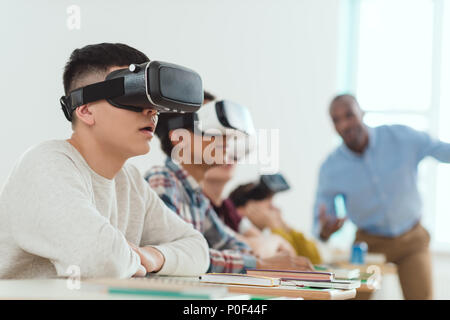 Side view of multicultural schoolchildren using virtual reality headsets and talking teacher standing behind Stock Photo