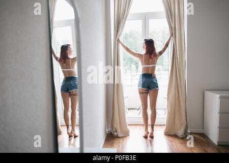 back view of young woman in bra and denim shorts opening curtains and reflecting in mirror Stock Photo