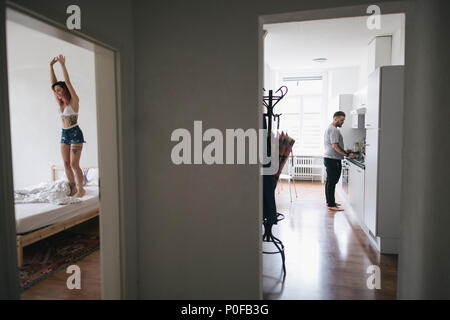 man cooking in kitchen while happy girlfriend jumping on bed in bedroom Stock Photo