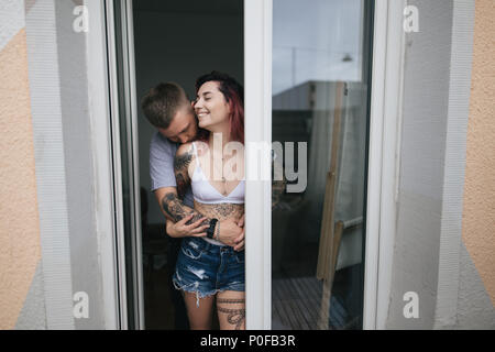 young man hugging and kissing beautiful smiling girlfriend in denim shorts and bra standing near window Stock Photo