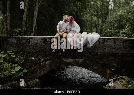 beautiful young wedding couple kissing while sitting on bridge above mountain river in Alps Stock Photo