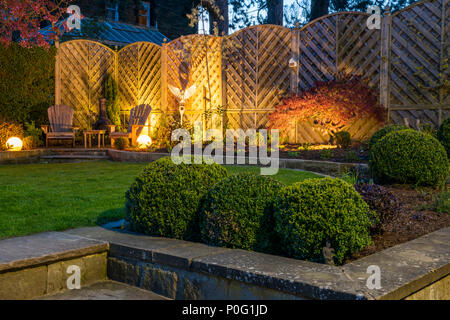 Corner of beautiful, landscaped, private garden with contemporary design, patio seating, box balls & lawn lit by globe lights - Yorkshire, England, UK Stock Photo