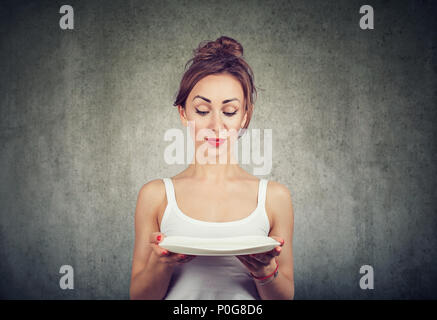 Young skinny girl holding empty plate and looking doubtful with diet restrictions standing on gray Stock Photo