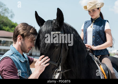 Handsome young man touching brown horse while woman in cowboy hat sitting on horseback Stock Photo