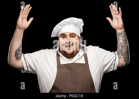 happy young chef in hat and apron gesturing with hands isolated on black Stock Photo