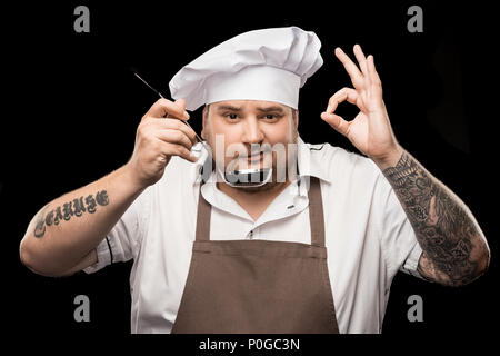 Smiling young chef holding scoop and showing ok sign isolated on black Stock Photo