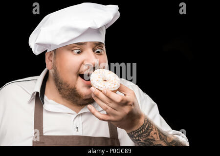 Excited chef in hat and apron eating doughnut isolated on black Stock Photo