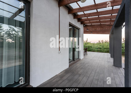 Side view of an open veranda in front of a modern forest cottage. Pine young forest under the sunset rays in the background. Stock Photo