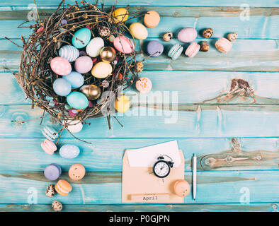 top view of colorful easter eggs in nest and envelope with alarm clock on blue wooden planks Stock Photo