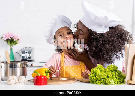 african american mother kissing her daughter in chef hat while cooking on kitchen Stock Photo
