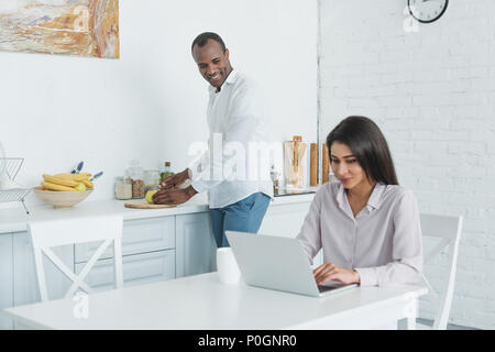 african american girlfriend using laptop and boyfriend cooking in morning Stock Photo