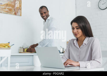 african american girlfriend using laptop and boyfriend cooking at kitchen Stock Photo