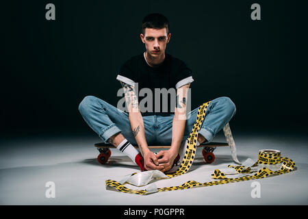 Hipster man sitting on longboard in police tape on dark background Stock Photo