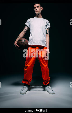 Man wearing prison uniform and cuffs holding basketball ball on dark background Stock Photo