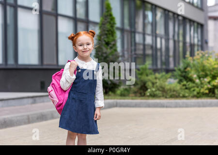 adorable red haired schoolgirl in spectacles holding backpack and smiling at camera Stock Photo