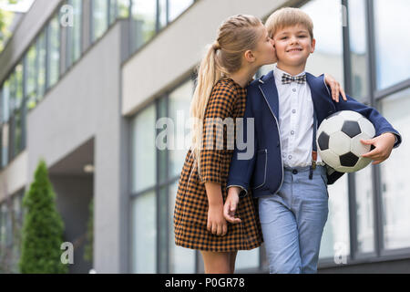 cute schoolgirl in dress kissing happy schoolboy with soccer ball Stock Photo