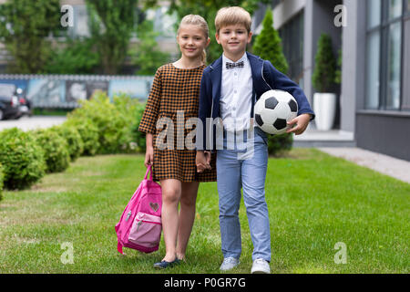 beautiful happy schoolboy and schoolgirl with backpack and soccer ball smiling at camera near school Stock Photo
