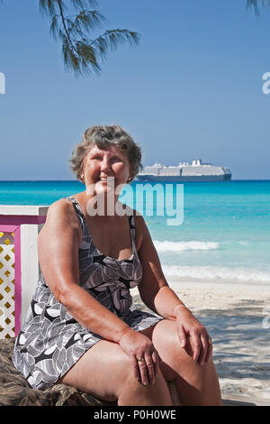 an older woman smiling and sunbathing on a beach with a cruise ship in  background Stock Photo