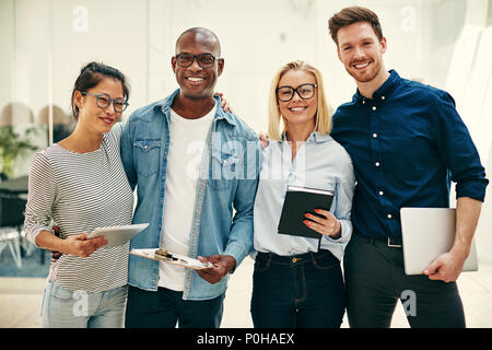 Group of diverse young businesspeople smiling confidently while standing arm in arm together in a bright modern office Stock Photo