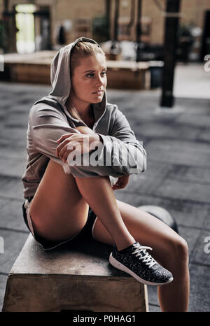 Fit young blonde woman wearing a hoodie sitting on a box before a workout in a gym Stock Photo