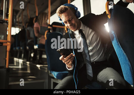 Young businessman smiling while sitting on a bus during his morning commute listening to music on headphones and using a smartphone Stock Photo