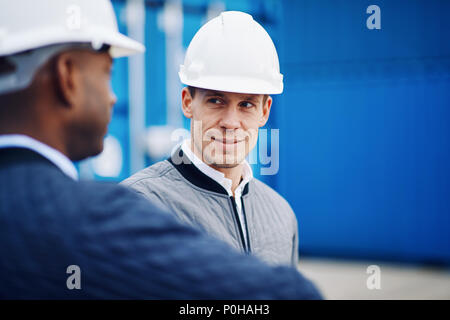 Smiling port manager wearing a hardhat standing by freight containers on a large commercial shipping dock talking with a colleague Stock Photo
