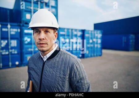 Port manager wearing a hardhat standing alone among freight containers on a large commercial shipping dock Stock Photo