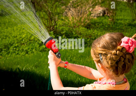Little child, adorable toddler girl, watering the plants, green lawn, from hose spray in the garden at the backyard of the house on a sunny summer day Stock Photo