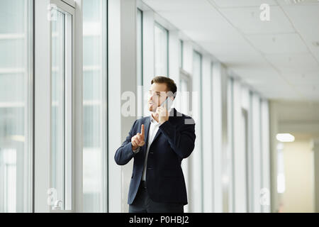 Young businessman standing near the window at office and talking on cell phone Stock Photo
