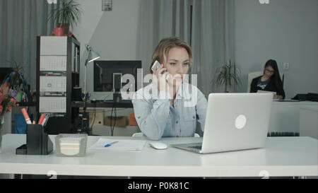 Young office secretary working on laptop and answering phone call Stock Photo